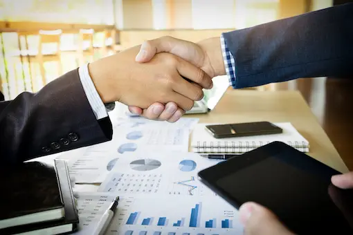 Two business professionals shaking hands over financial charts and data reports to finalize a procurement and acquisition agreement