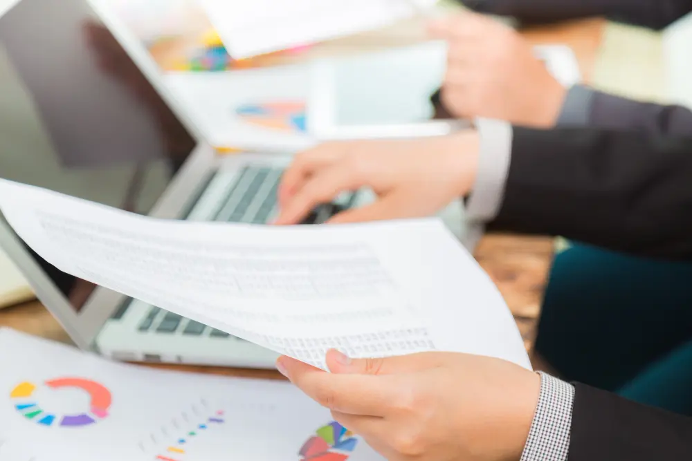 A close-up of a business professional holding printed reports while working on a laptop, illustrating the detailed documentation and review phase of a Project Procurement Management Plan.