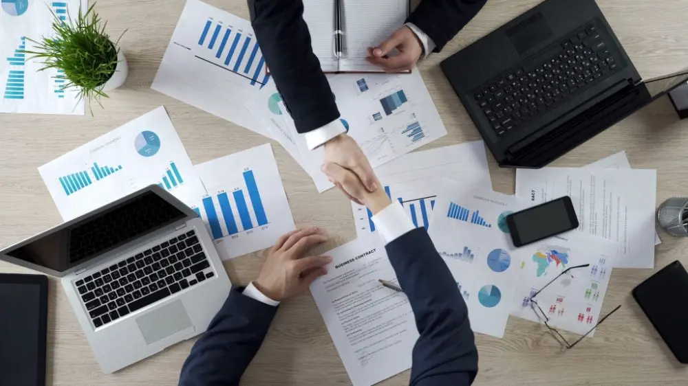 Business professionals shaking hands over financial reports, charts, and laptops during a corporate partnership meeting.