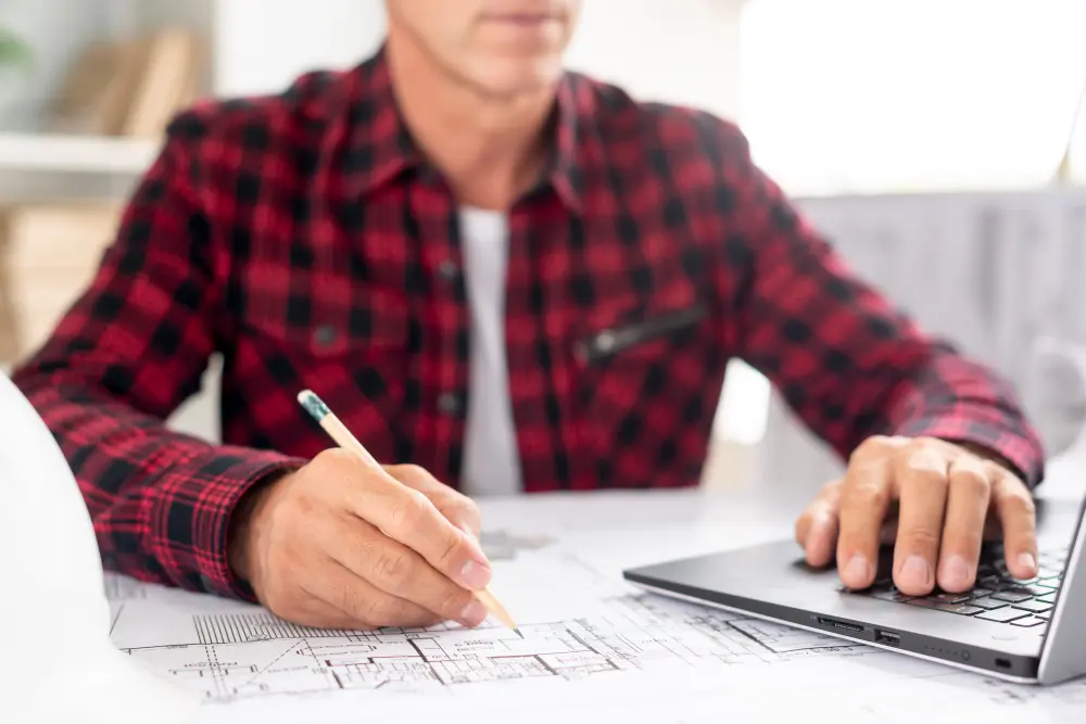 Person reviewing architectural floor plans while using a laptop and pencil for construction project planning